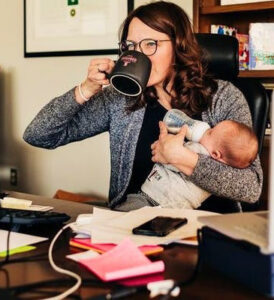 Woman behind a desk drinking a coffee while holding a baby drinking from a bottle